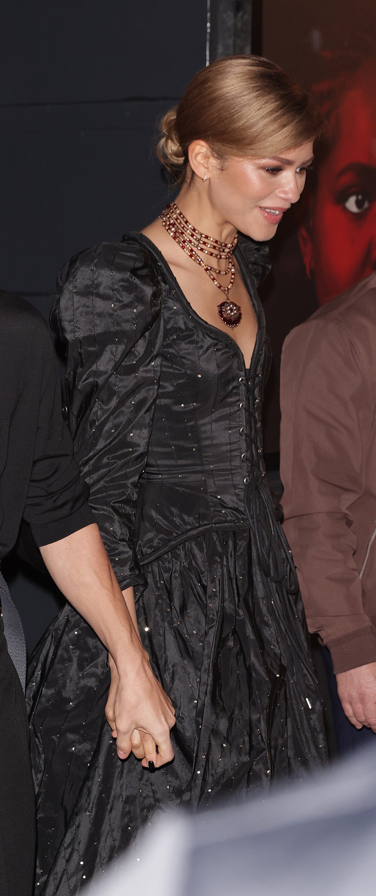 Tom Holland and Zendaya holding hands, dressed in stylish black outfits, attending an event. Zendaya wears a black dress with puffed sleeves, and Tom wears a black shirt with pants