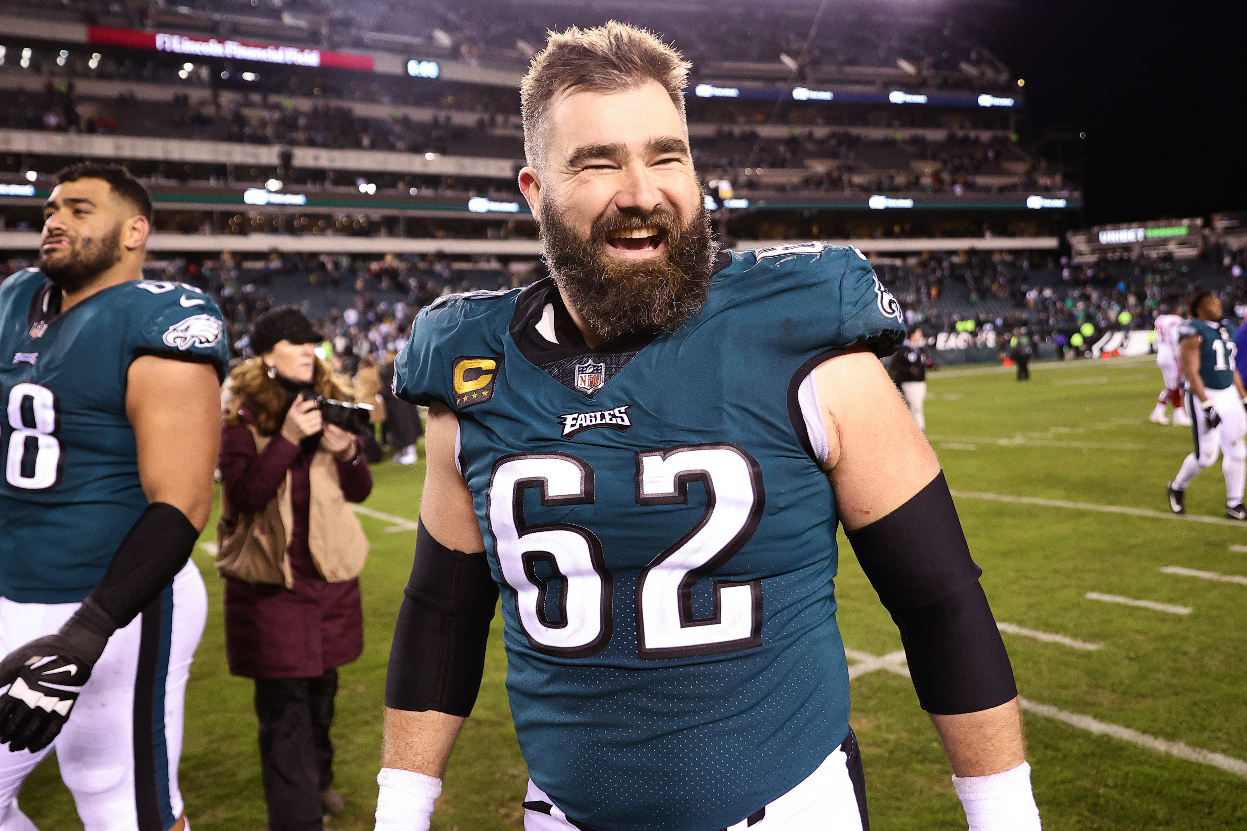 Jason Kelce from the Philadelphia Eagles smiles widely on the football field post-game, wearing his number 62 jersey. Another player walks in the background