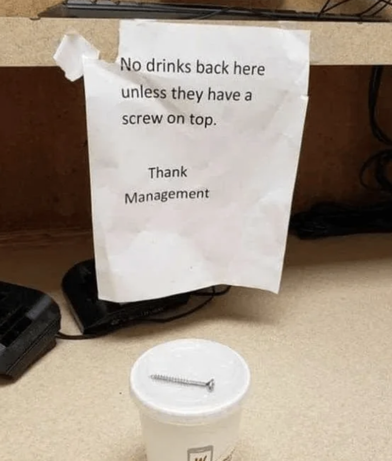 A notice reads, "No drinks back here unless they have a screw on top. Thank Management." Below the notice, a cup with a screw on its lid is placed on the table