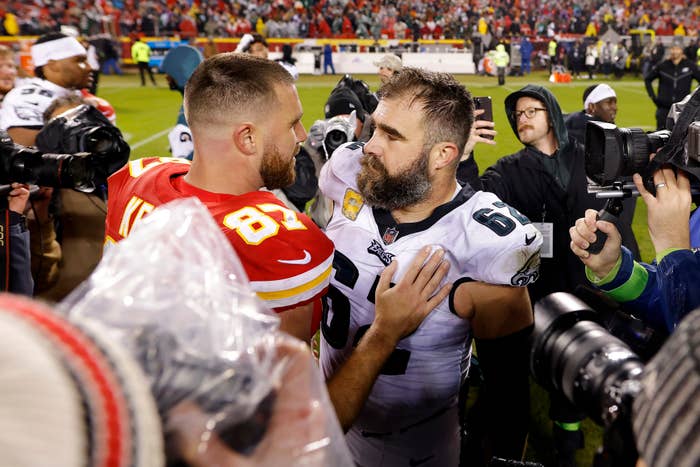 Travis Kelce and Jason Kelce, in football uniforms, share a moment on the field after a game surrounded by photographers and fans