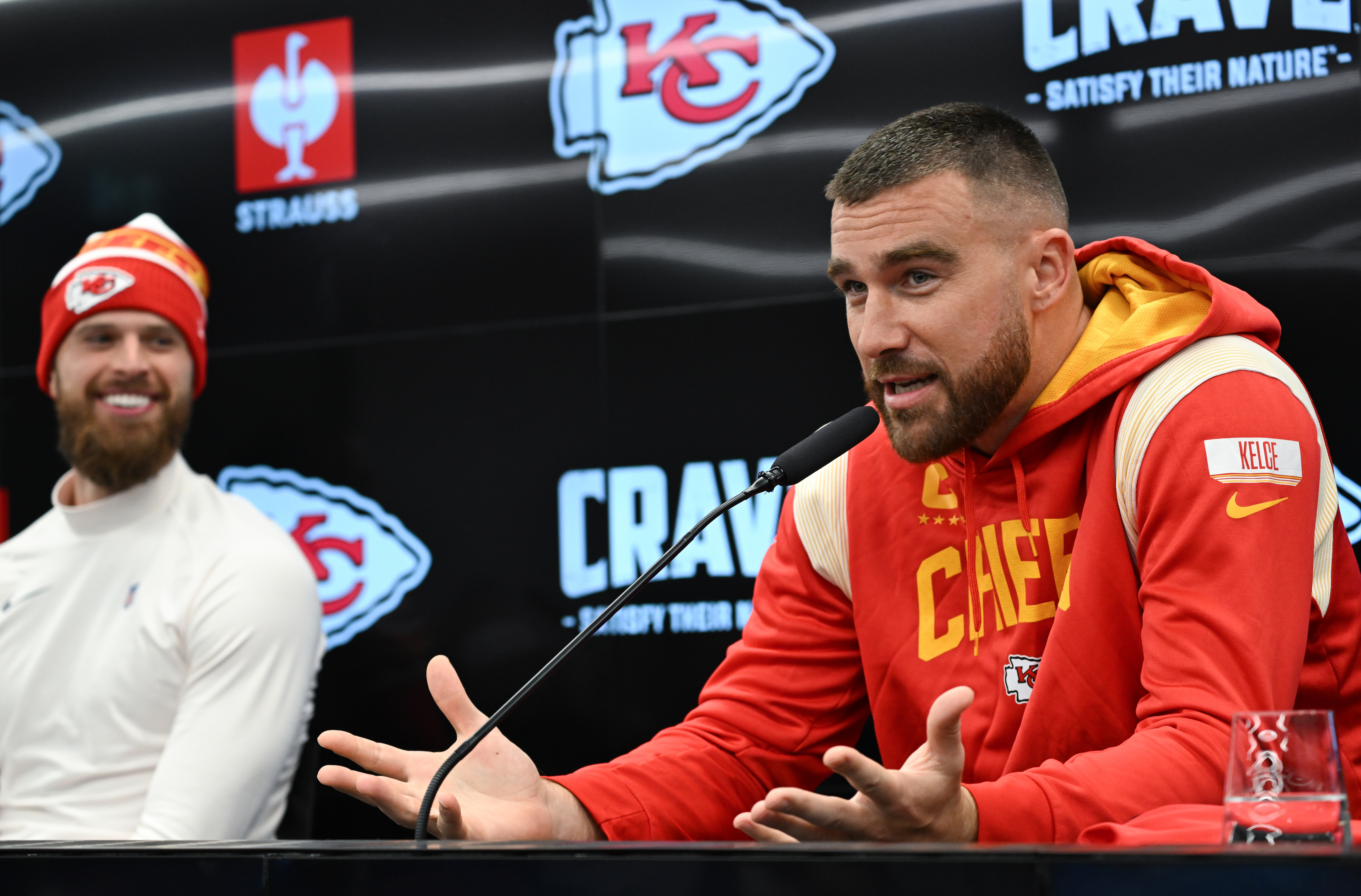 Kansas City Chiefs player Travis Kelce speaks at a press conference, gesturing with his hands, while Patrick Mahomes sits smiling nearby