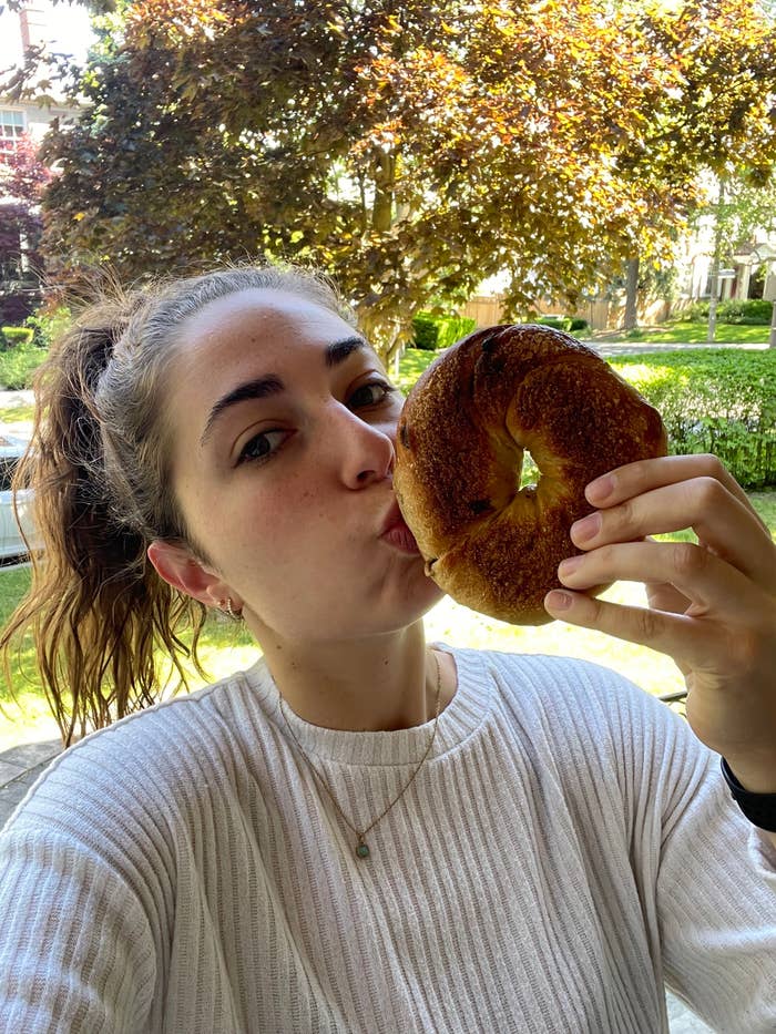 A woman holds a bagel close to her face while smiling in an outdoor setting with trees and buildings in the background