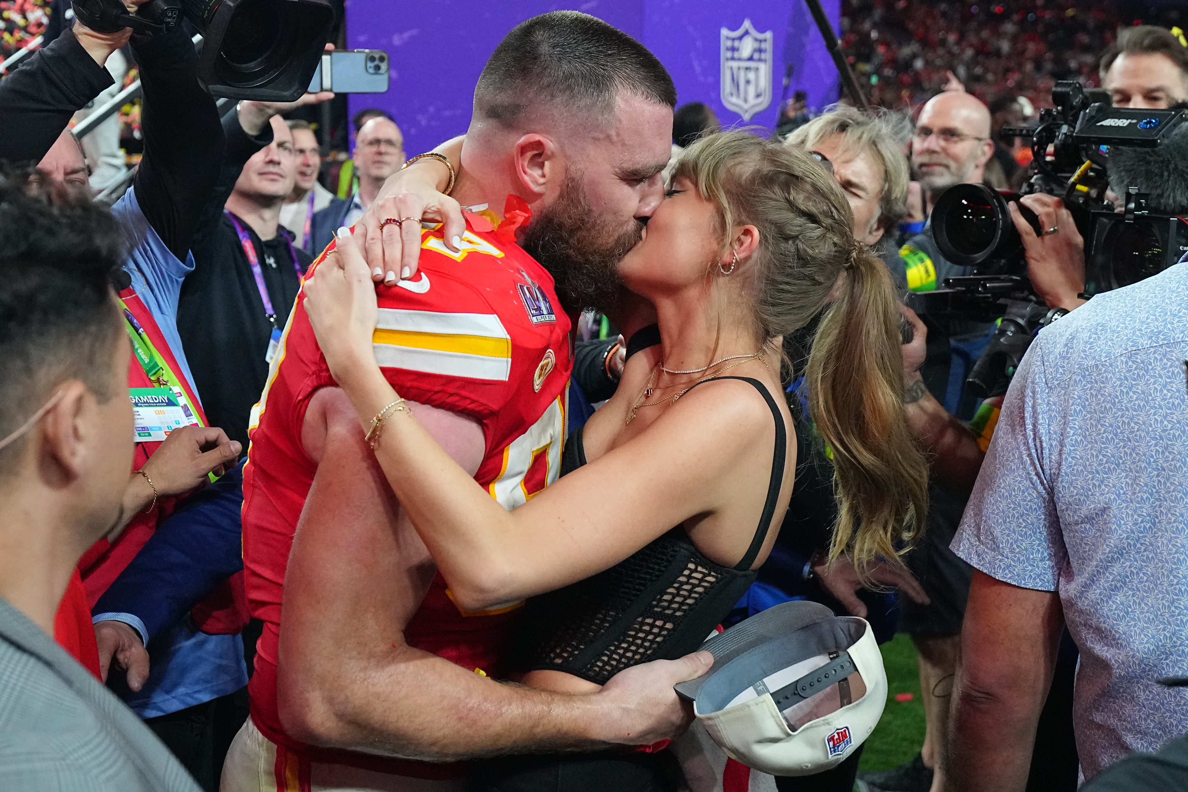 Travis Kelce and Taylor Swift share a kiss on the sidelines at an NFL game, surrounded by photographers and onlookers