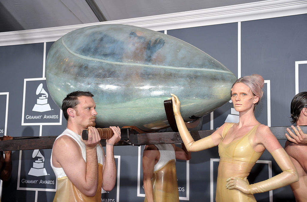 Artists at the Grammy Awards carrying an art piece resembling a large, glossy pod. They are wearing tight, glossy outfits, accentuating the futuristic theme