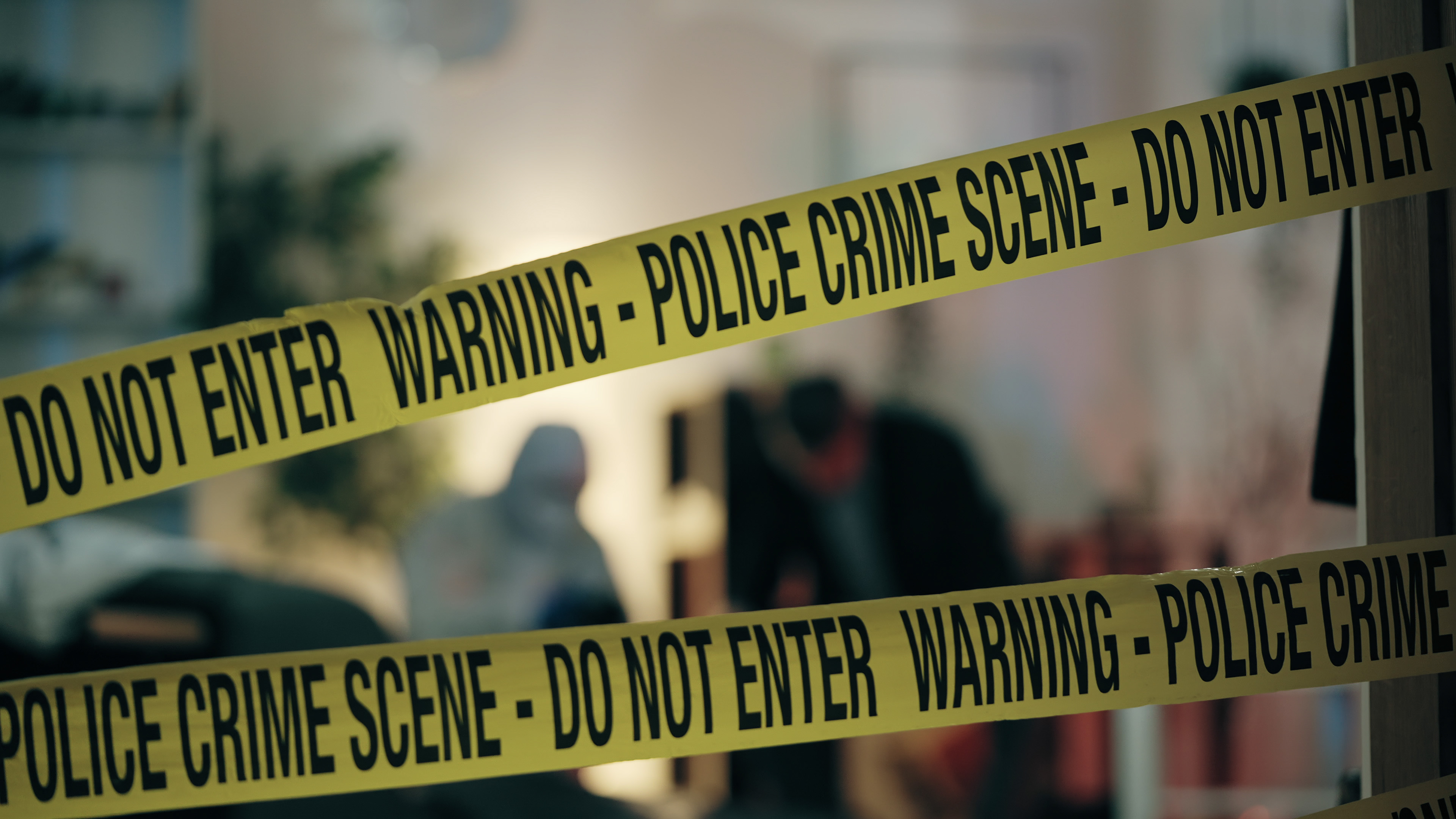 Police crime scene tape with "Warning - Do Not Enter" barriers blocking entrance to a blurred indoor area with people in the background