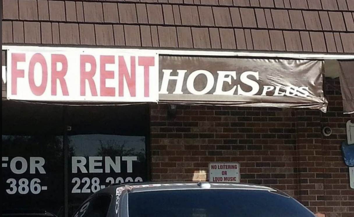 A sign on a brick building reads "FOR RENT" with a torn banner next to it that appears to spell "HOES". A car is parked in front