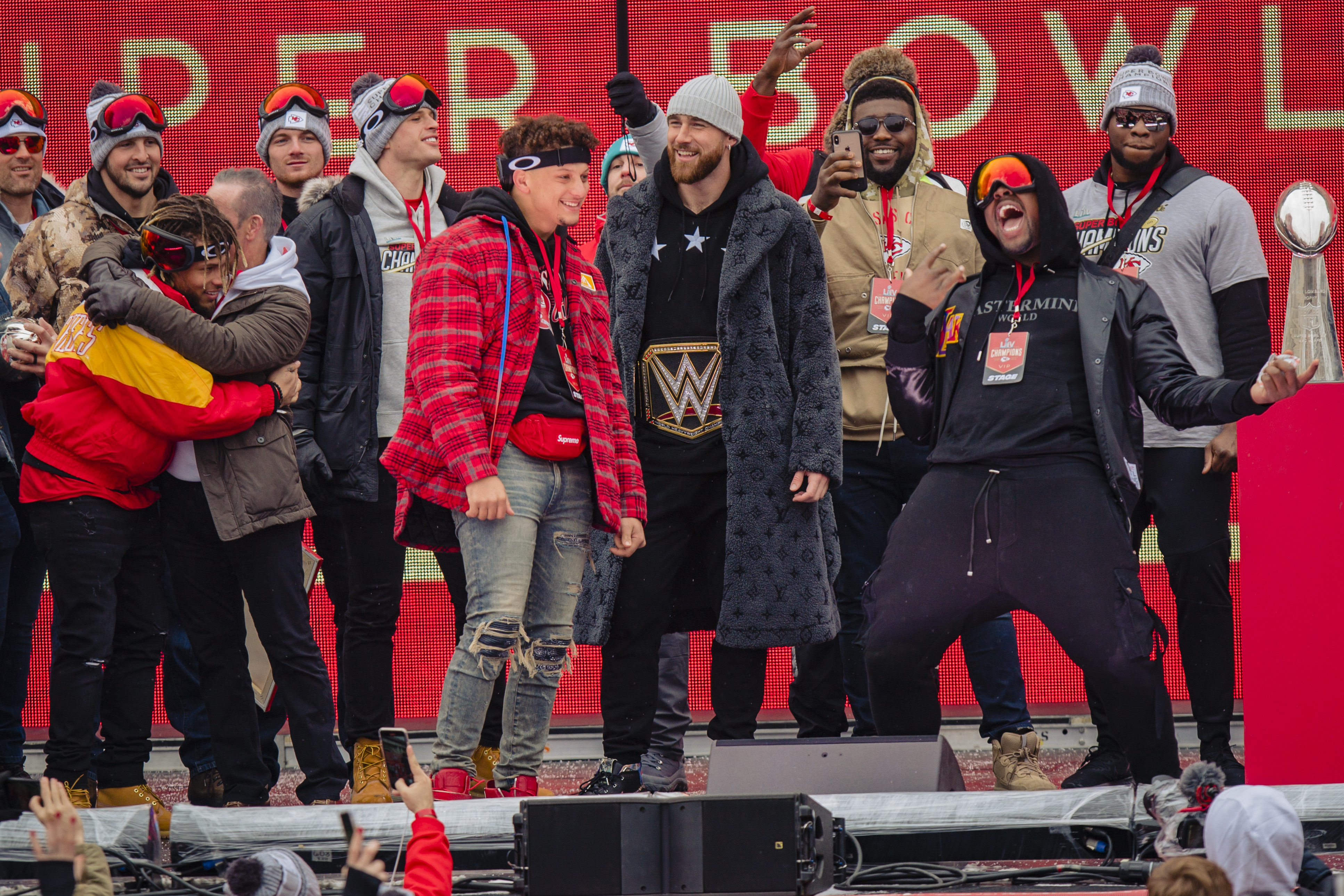 Group of NFL players, including Travis Kelce and Patrick Mahomes, celebrate on stage with a WWE belt and a Super Bowl trophy, dressed in casual and team apparel