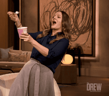 Drew Barrymore enthusiastically eats ice cream from a tub with a spoon while on the set of "The Drew Barrymore Show."