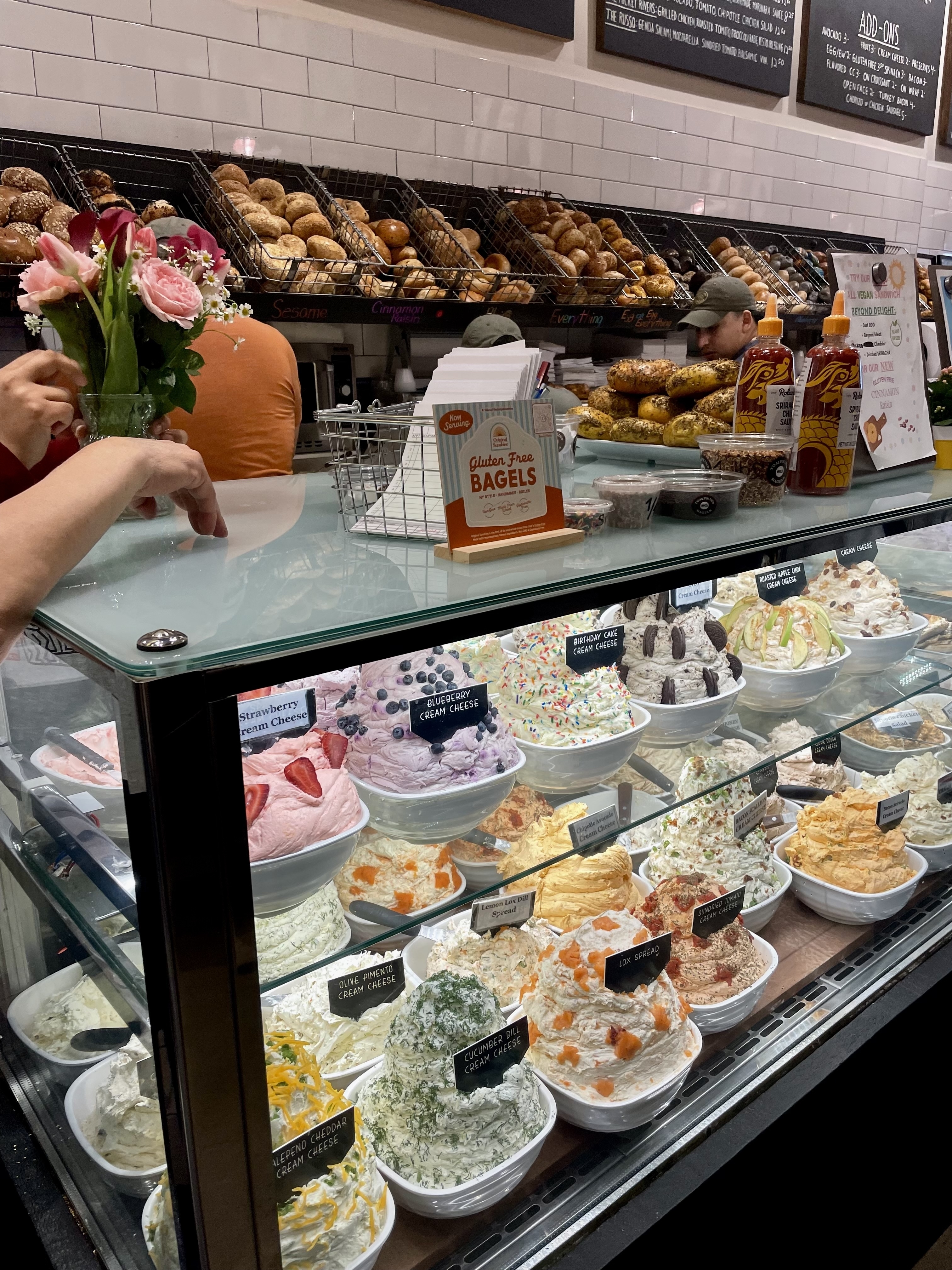 A deli counter with various flavors of cream cheese displayed in bowls, a flower bouquet, bagels, and a selection of bagel spreads. A person is arranging flowers