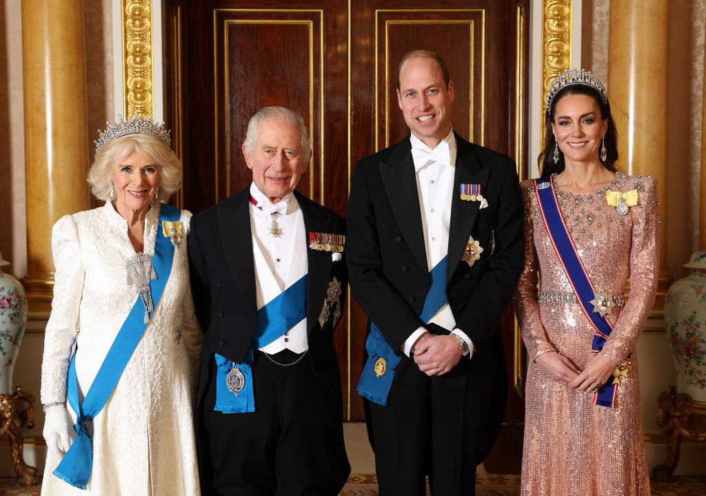 Camilla, King Charles III, Prince William, and Kate Middleton standing in formal attire with sashes and medals, posing for a photo indoors