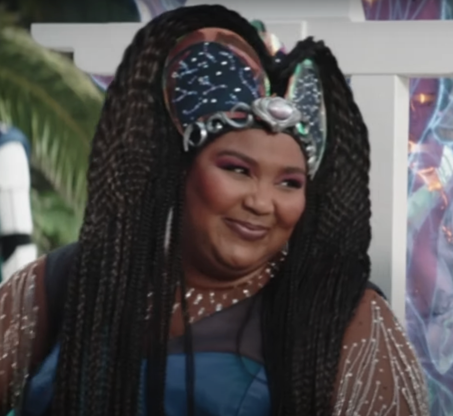Lizzo wearing an elaborate, futuristic headpiece with long braids, smiles on a set with futuristic and colorful background elements in a scene