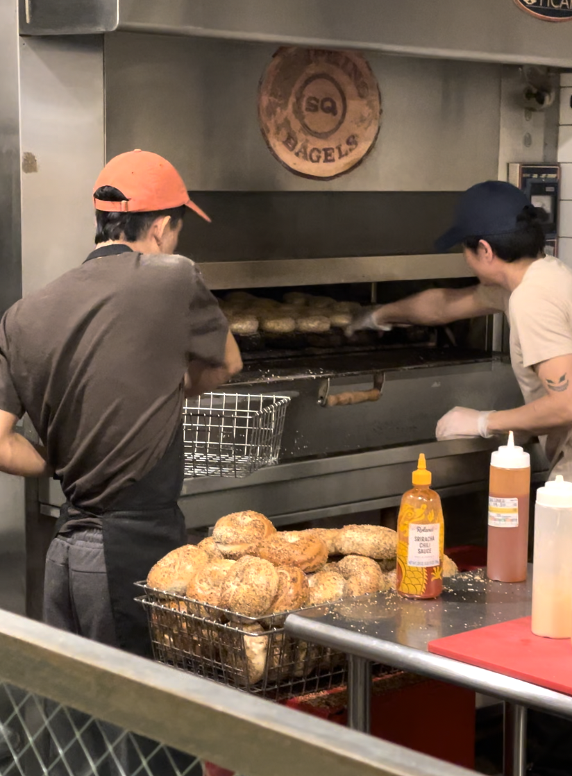 Two people working at a bakery, pulling bagels out of an oven. Baskets of bagels and condiment bottles are visible on the counter