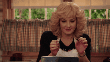 A woman with blonde curly hair smiles and looks at a toaster while placing something inside. She is dressed in an apron and kitchen setting