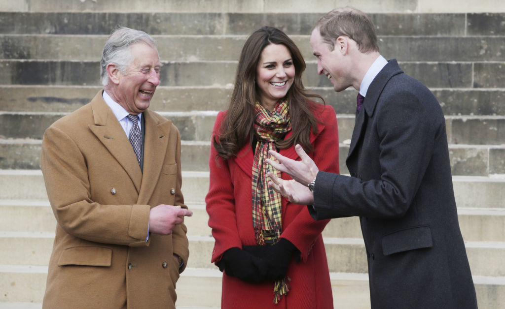 Charles, Kate, and William stand outdoors, engaged in conversation. Charles and William are dressed in suits and coats, while Kate wears a red coat and scarf, smiling
