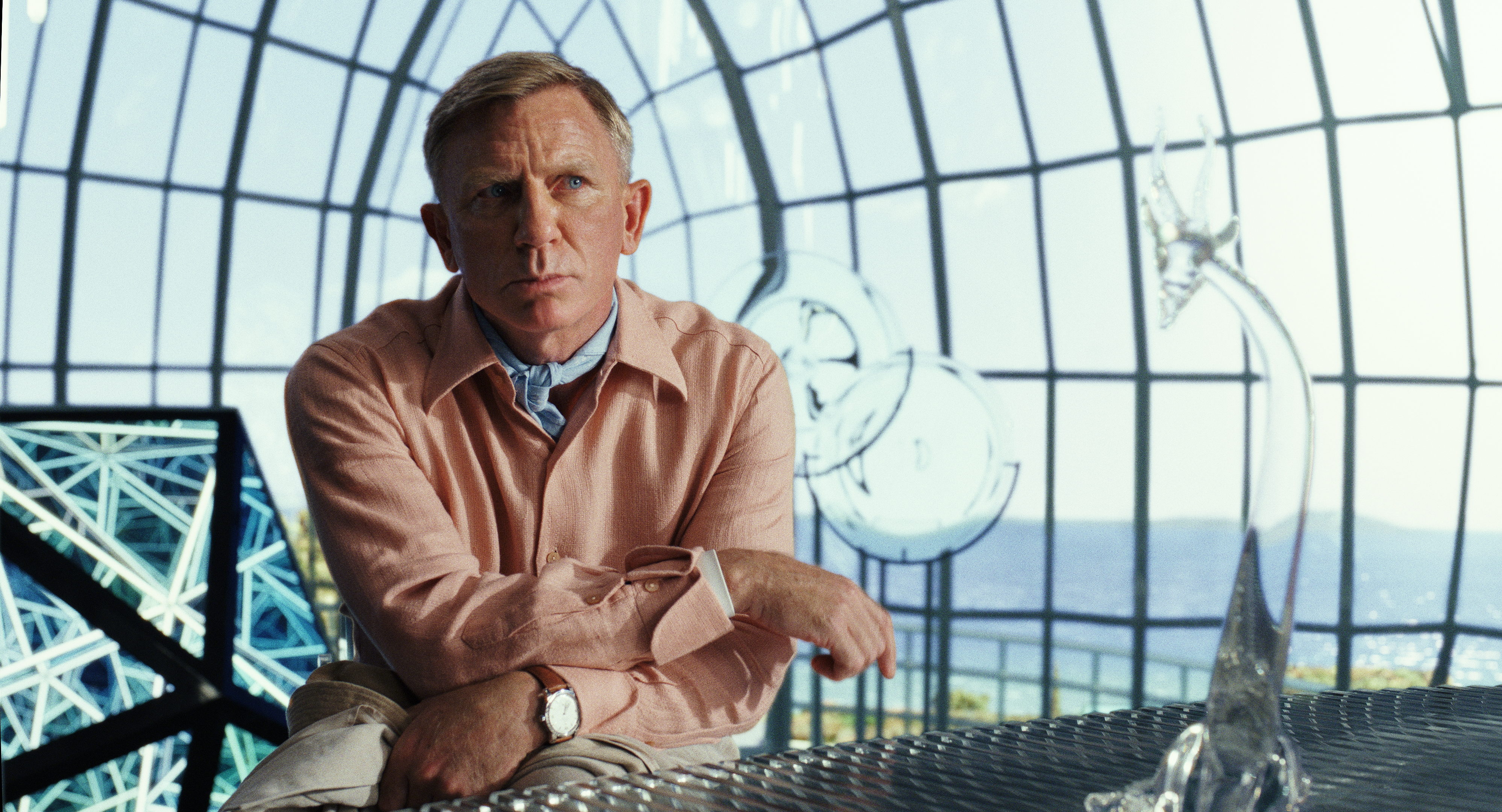 Daniel Craig, wearing a shirt with a scarf, sits thoughtfully at a glass table in front of a large, modern, geometric window with a scenic view
