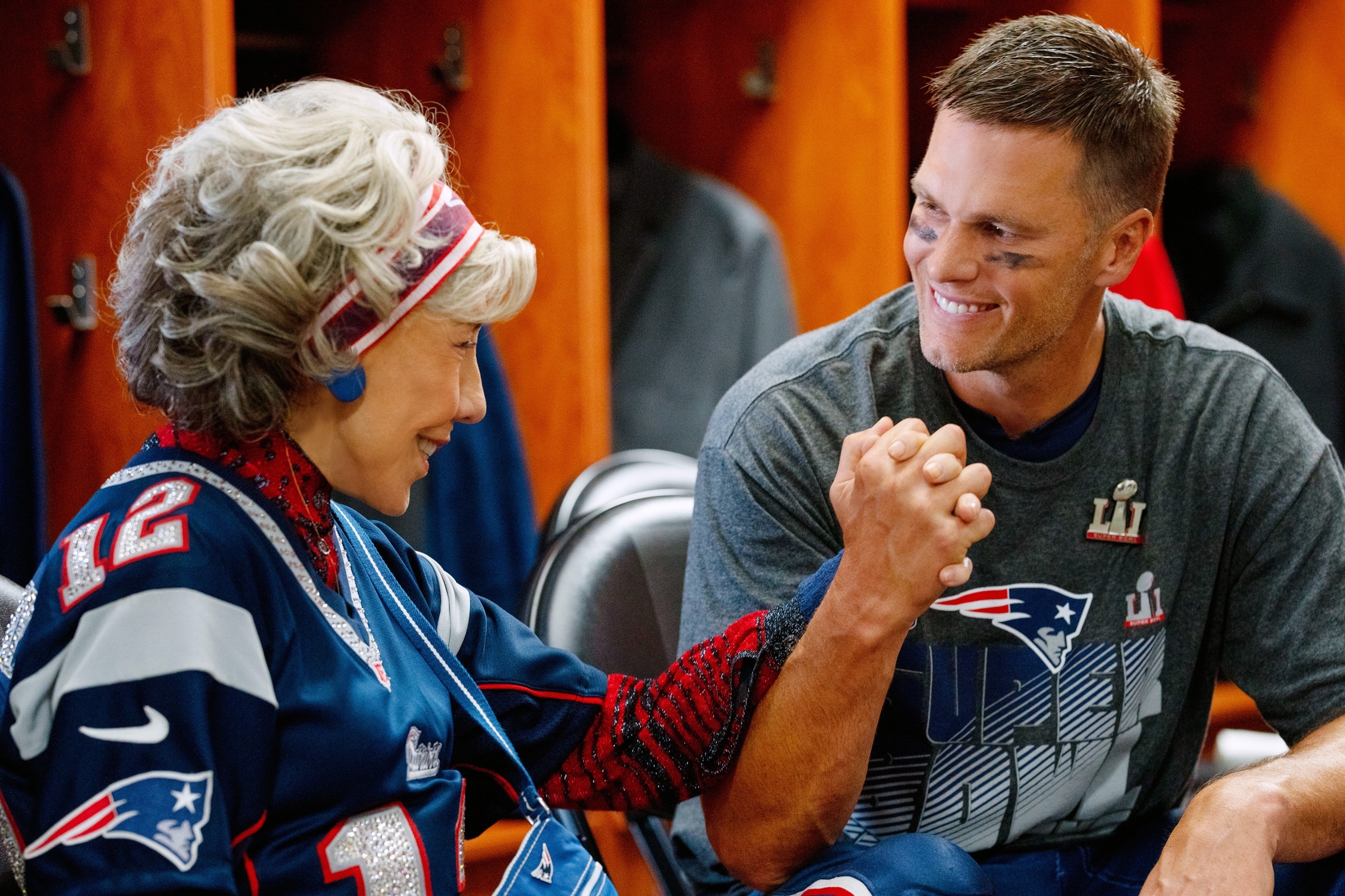 Lily Tomlin and Tom Brady smile and clasp hands in a locker room. Lily wears a Patriots jersey, and Tom is in a casual Patriots shirt, in a scene