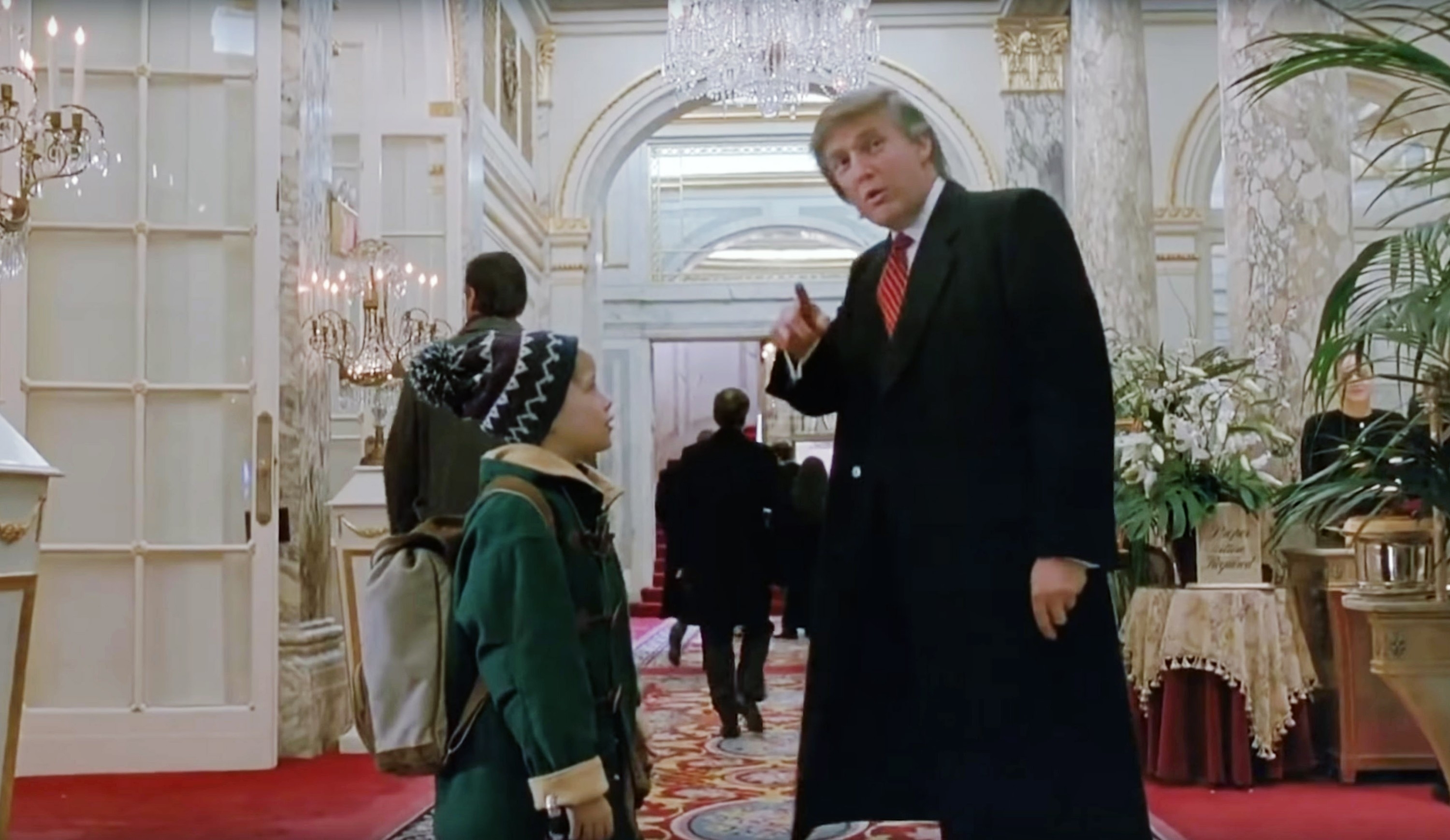 Donald Trump, wearing a suit and tie, points while speaking to a child bundled in winter attire inside an ornate building with marble columns and chandeliers in a scene