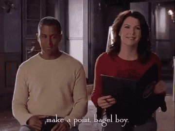 Lauren Graham in a red top holding a menu, sitting next to Yanic Truesdale in a light sweater, with text overlay: "make a point, bagel boy."