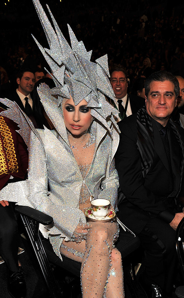 Lady Gaga sits in a dramatic, angular outfit holding a teacup, with several people around her at a formal event