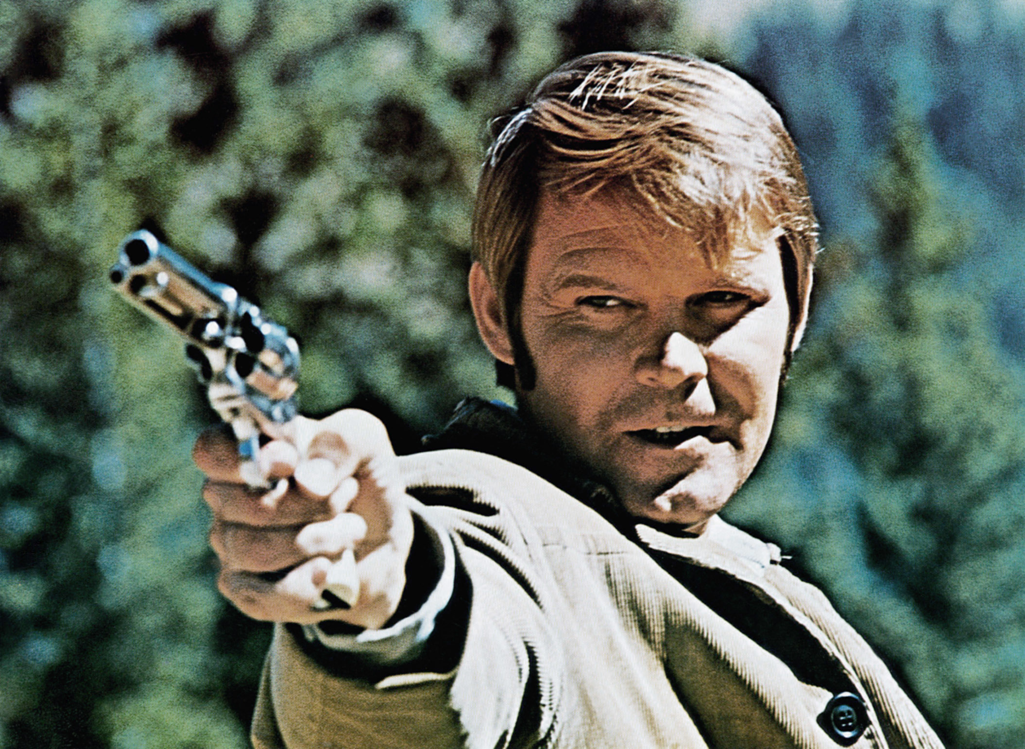 Glen Campbell points a revolver at the camera in a dramatic scene. He appears intense, set against a blurred outdoor background