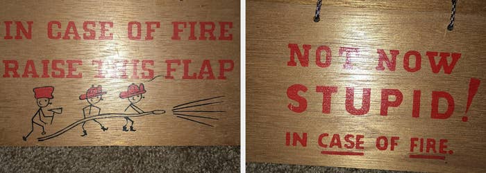 Two wooden signs show a fire emergency warning. The first reads "IN CASE OF FIRE RAISE THIS FLAP", and the second says "NOT NOW STUPID! IN CASE OF FIRE."