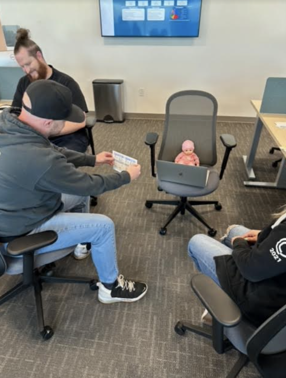 Four people are seated in an office. Three adults surround the doll seated on a swivel chair with a laptop