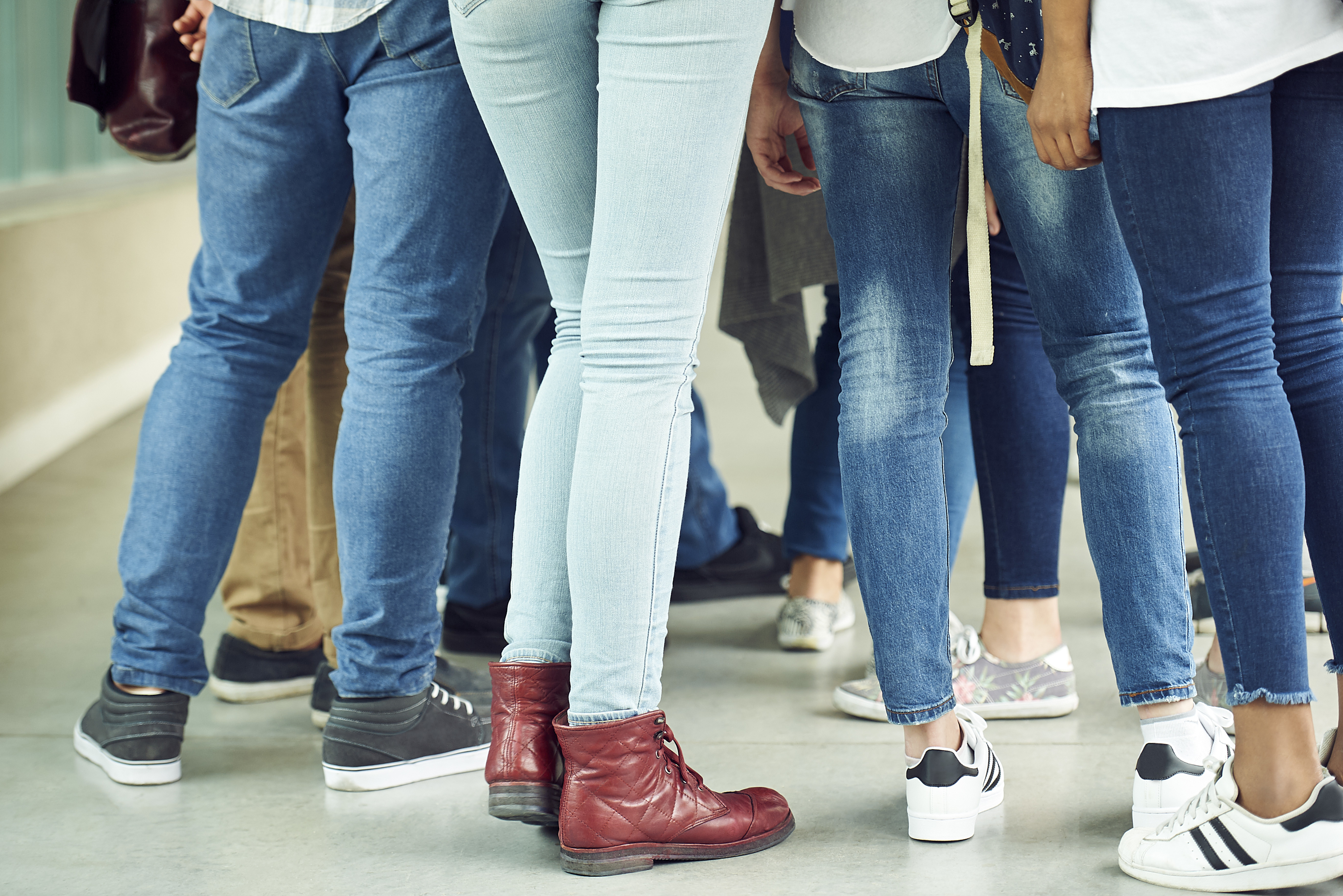 A group of people, mostly wearing jeans and casual shoes, stand close together. The focus is on their lower bodies and footwear