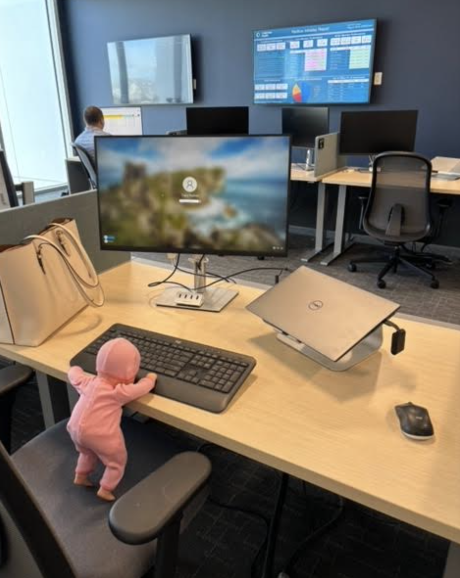 A baby doll is propped up against a keyboard on a desk in an office setting. Multiple desks and monitors are visible in the background