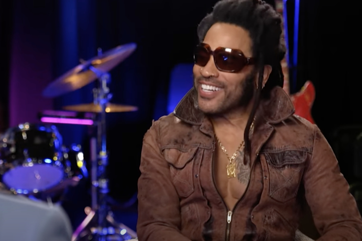 Lenny Kravitz, wearing sunglasses and a brown leather jacket, smiles during an interview in a studio with a drum set and guitar in the background