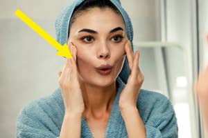 A woman in a blue towel head wrap and robe applies skincare to her face in front of a mirror