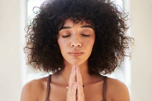 A woman with curly hair closes her eyes and brings her hands together in a prayer or meditation pose