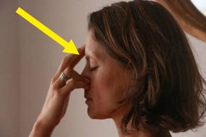 A woman with shoulder-length hair is performing a breathing exercise, pressing her fingers against her nose. A yellow arrow points to her face