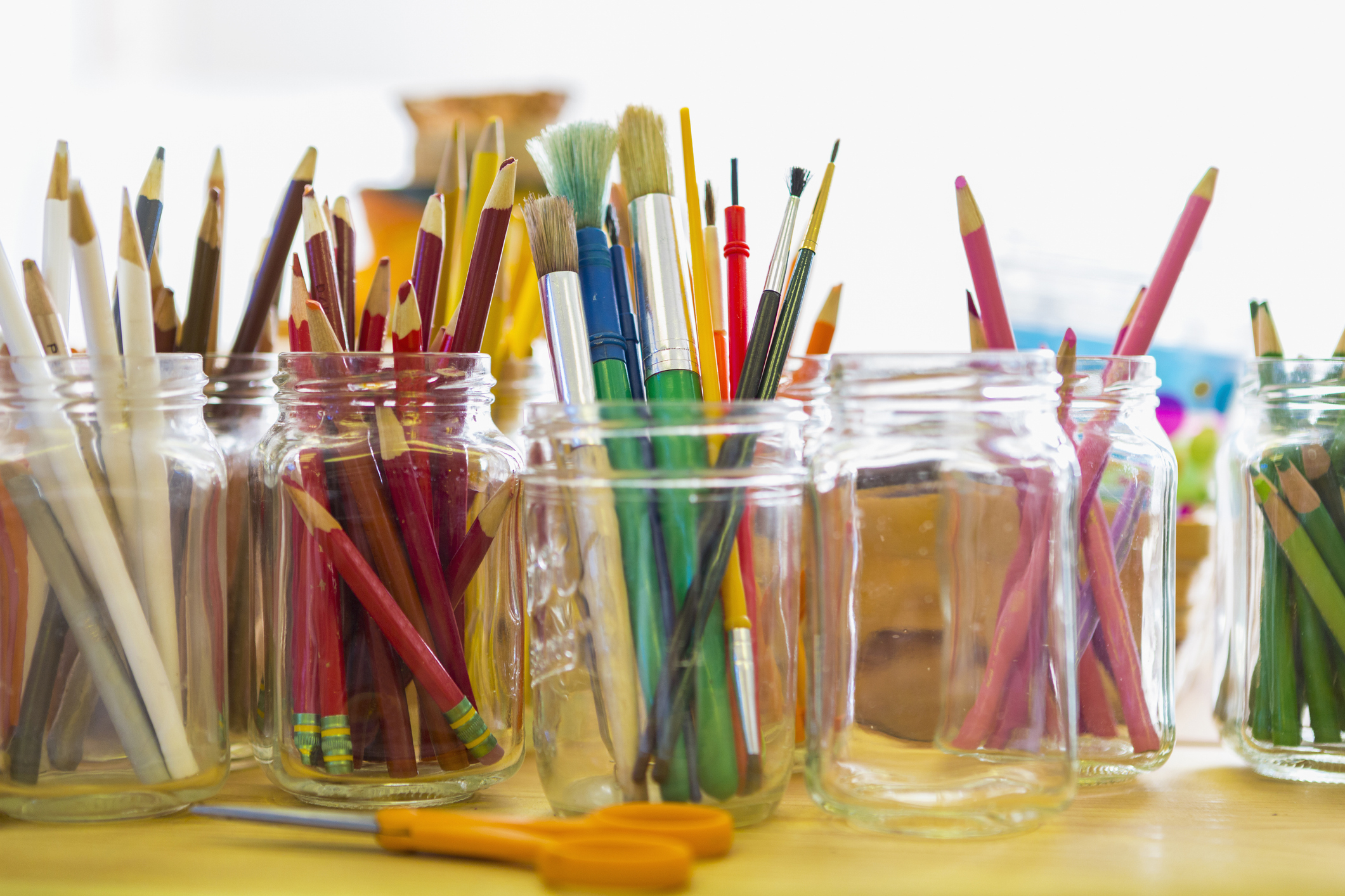 Mason jars filled with various art supplies like pencils, paintbrushes, and markers on a table; orange scissors are seen in the foreground