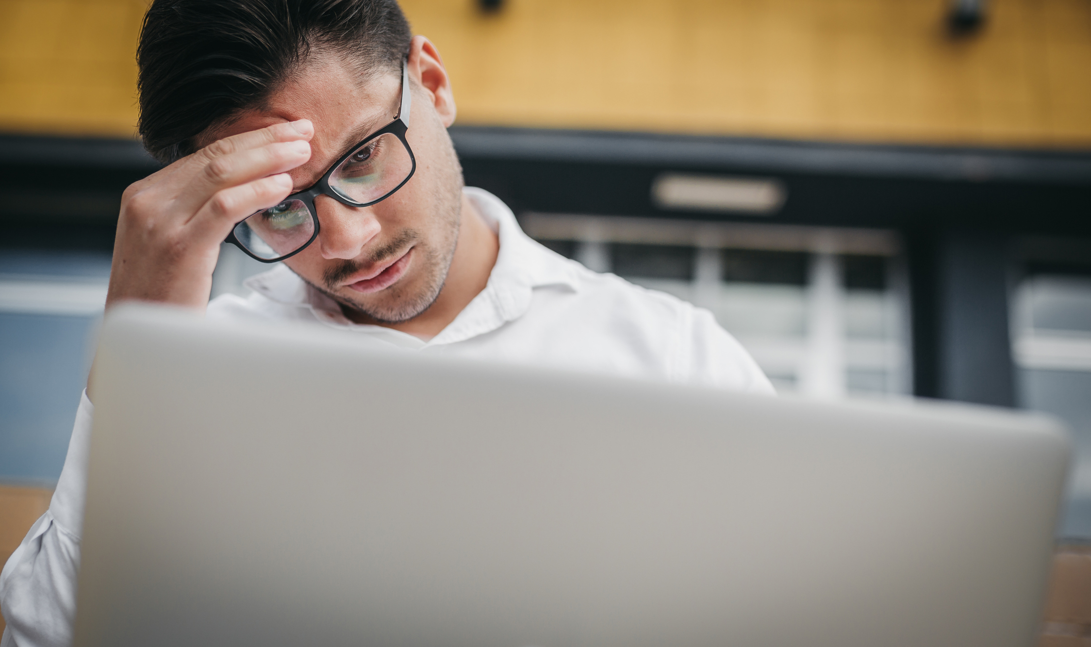 Man with glasses looks stressed, holding his forehead while looking at a laptop screen