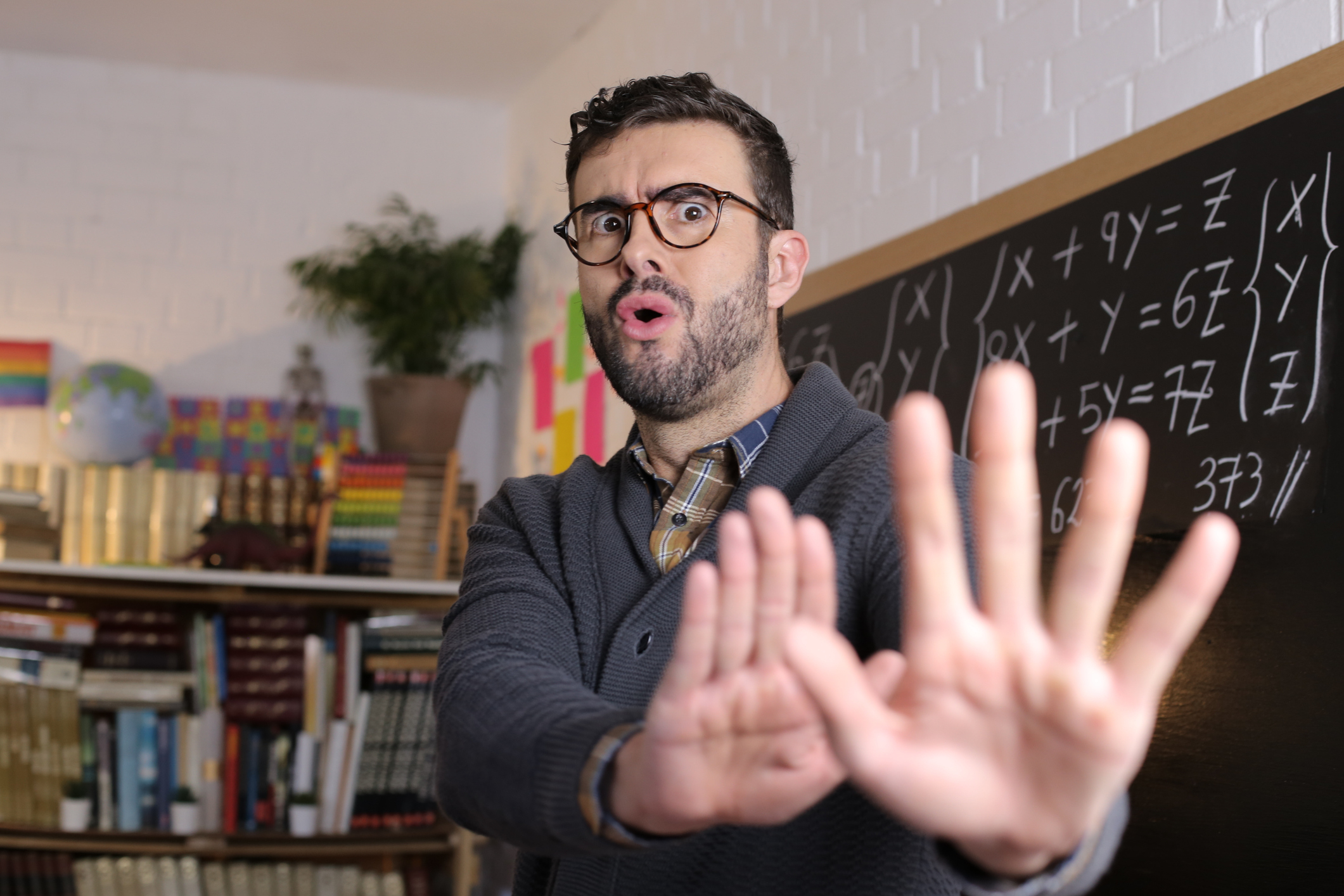 Teacher in a cozy classroom, appearing surprised or concerned, with hands raised in a stopping motion in front of a blackboard with math equations