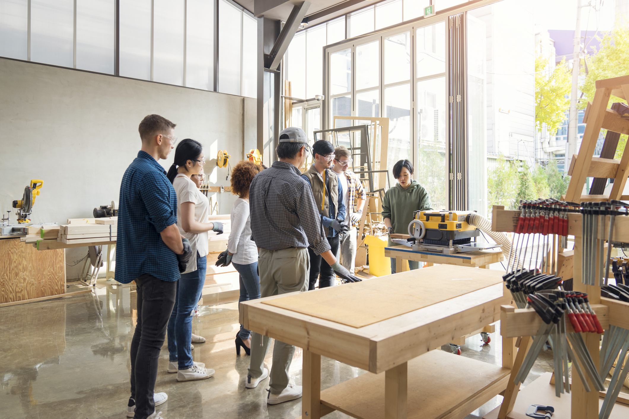 A group of adults and teenagers gather around a woodworking table, attentively watching a demonstration in a bright, modern workshop