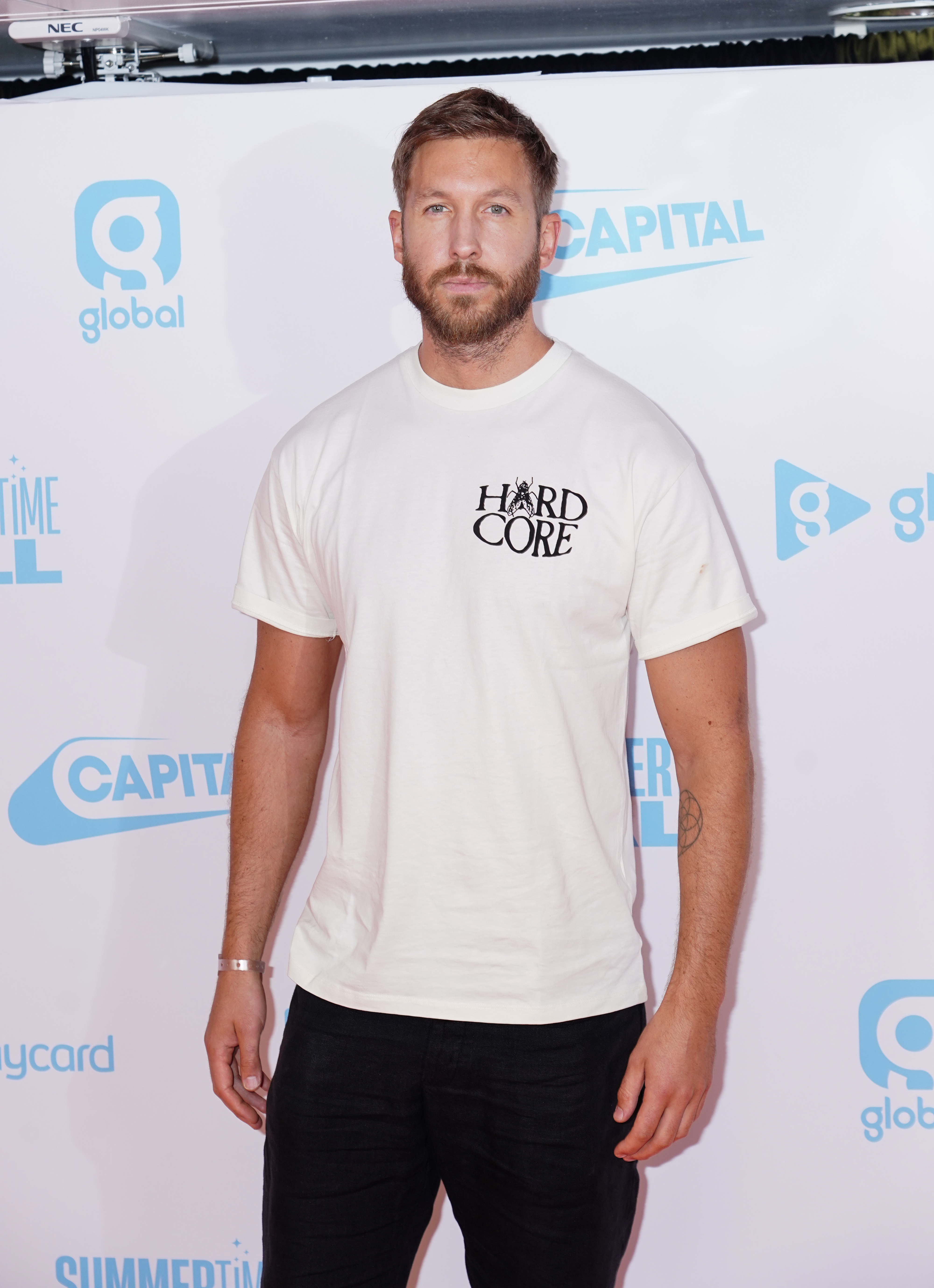 Calvin Harris on the red carpet wearing a white t-shirt with "HARDCORE" printed on it and black pants at Global's Summertime Ball event