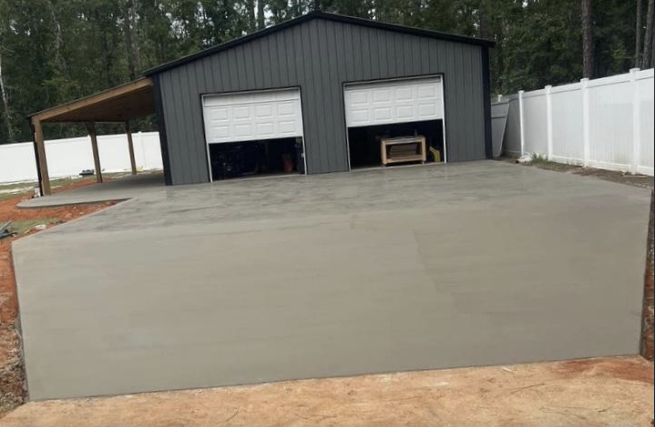 A garage with two white doors and a fresh concrete slab in front. The structure is set in a wooded area with a white fence around the property. No people are present