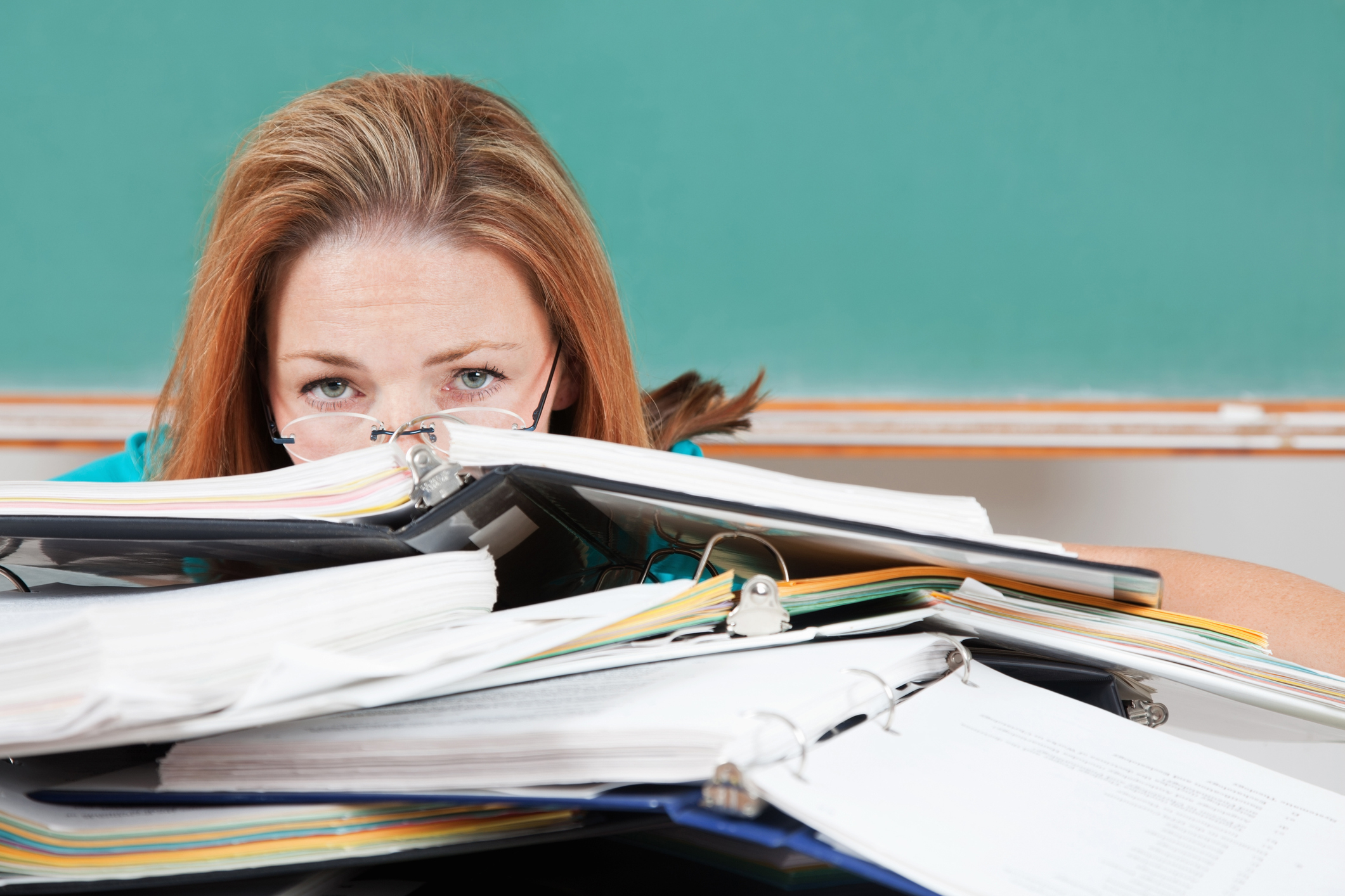 A woman, partially hidden behind several open binders and stacks of papers on a desk, appears overwhelmed
