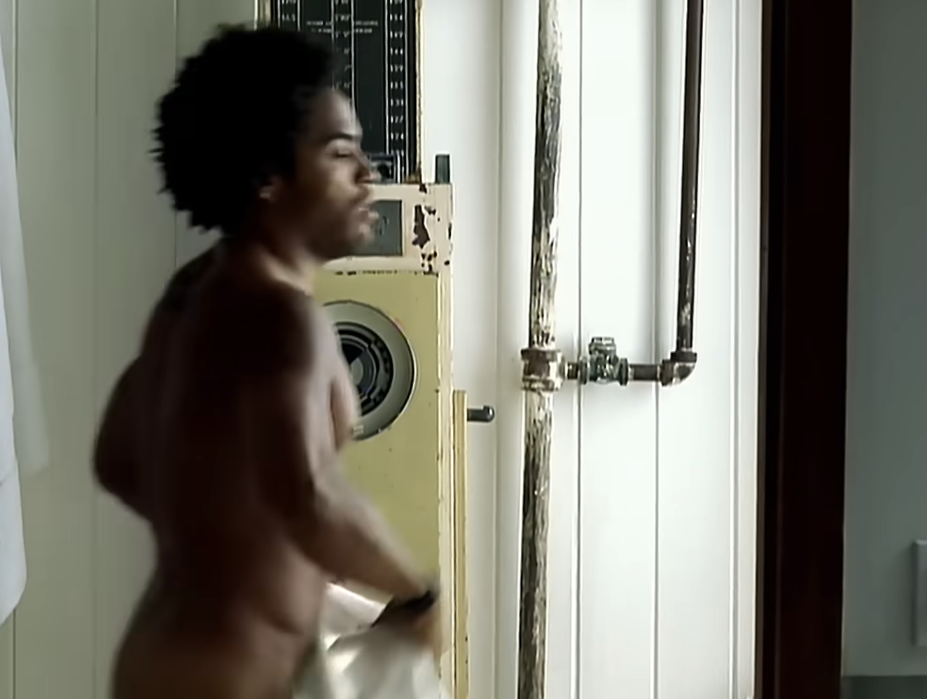 Donald Glover in a bare room with industrial pipes behind him, holding a towel