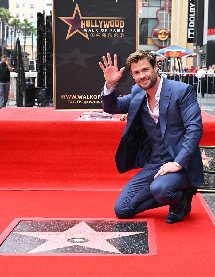 Chris Hemsworth kneels and waves next to his star on the Hollywood Walk of Fame, wearing a light suit and shirt without a tie