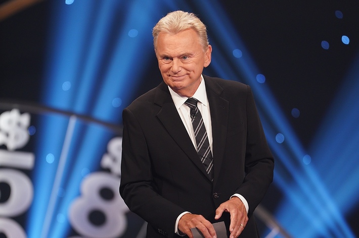 Pat Sajak, the host of "Wheel of Fortune," in a suit and tie, standing on the show's illuminated stage