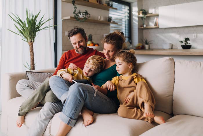 A family of four, with parents and two young children, sit closely on a sofa reading a book together in a cozy living room