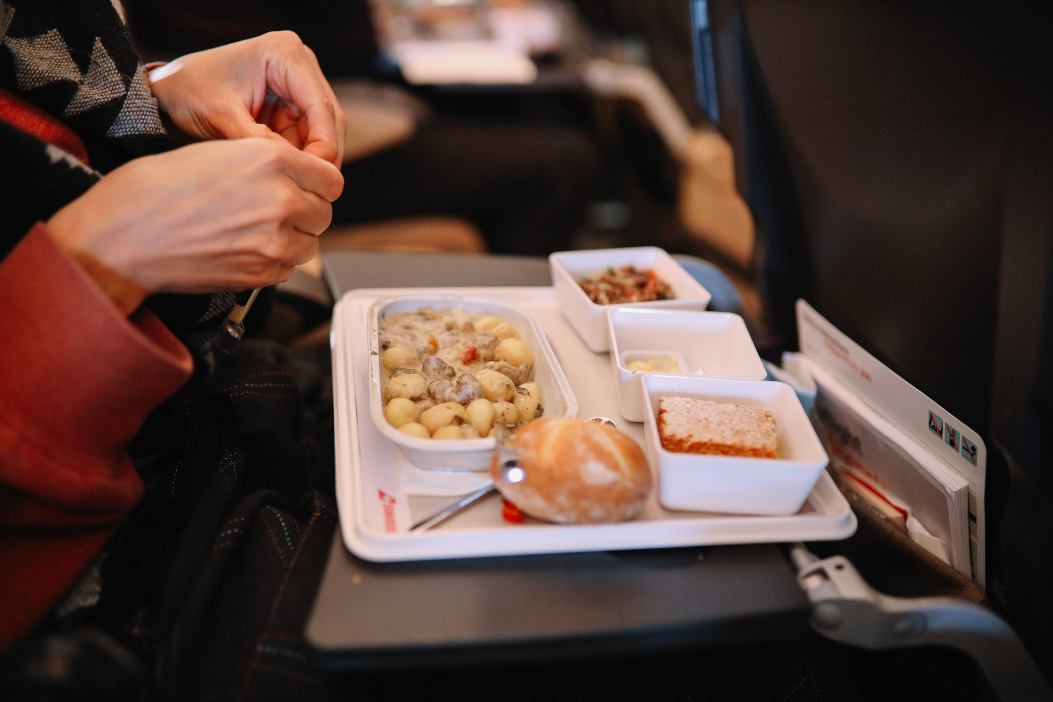 A person is preparing to eat an airline meal consisting of pasta, bread roll, dessert, and side dishes on a tray in an airplane