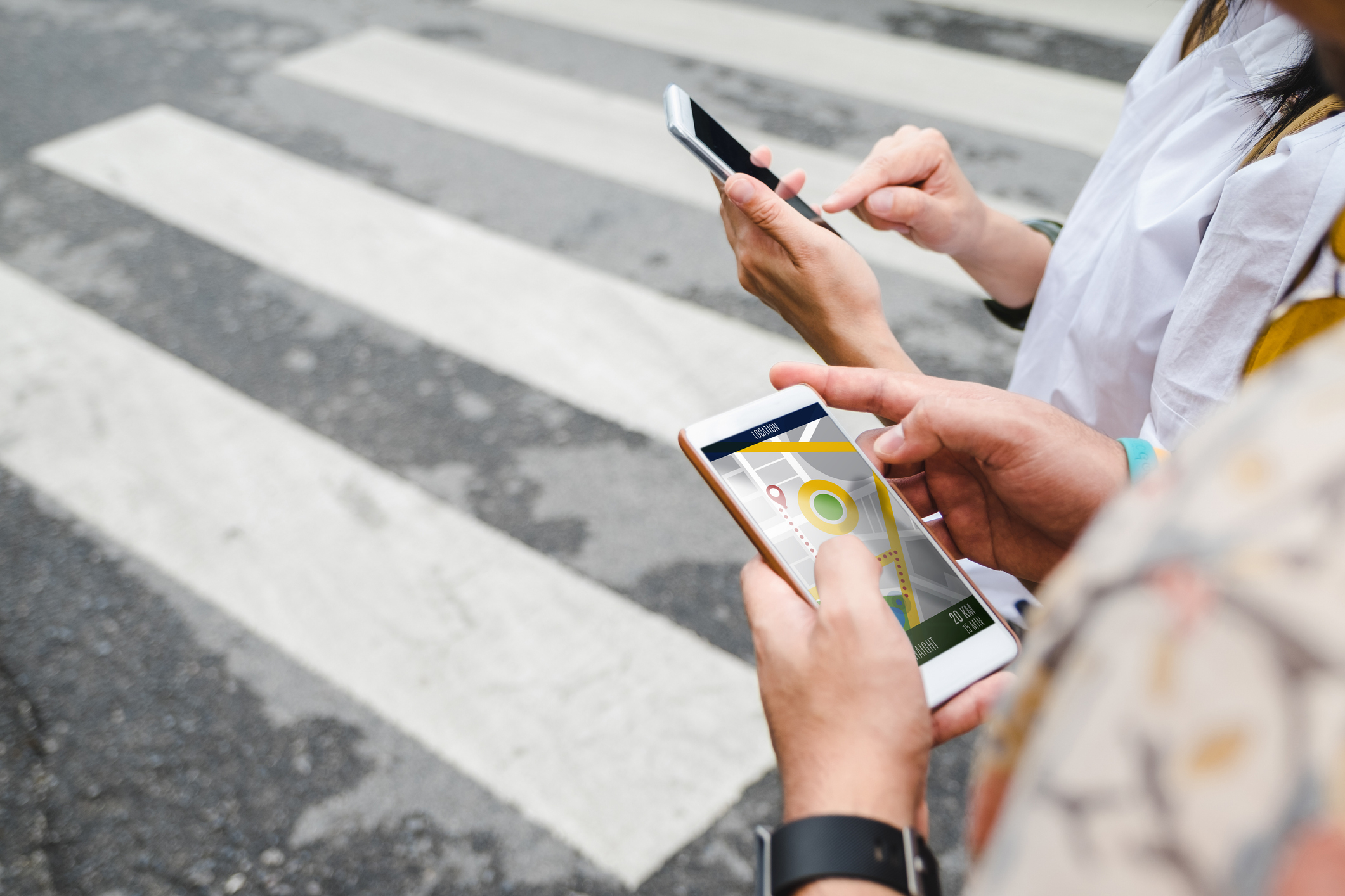 Three people using smartphones while crossing a street with a pedestrian crosswalk. One phone displays a map application