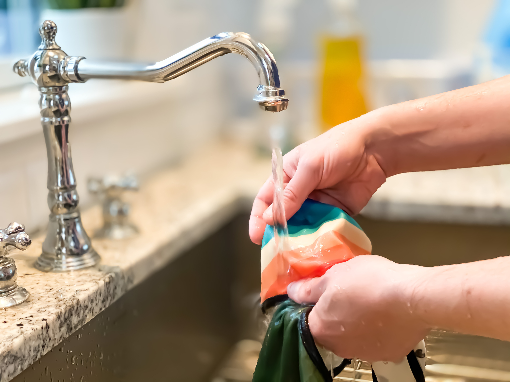 A person washes a multicolored bowl under a kitchen sink faucet