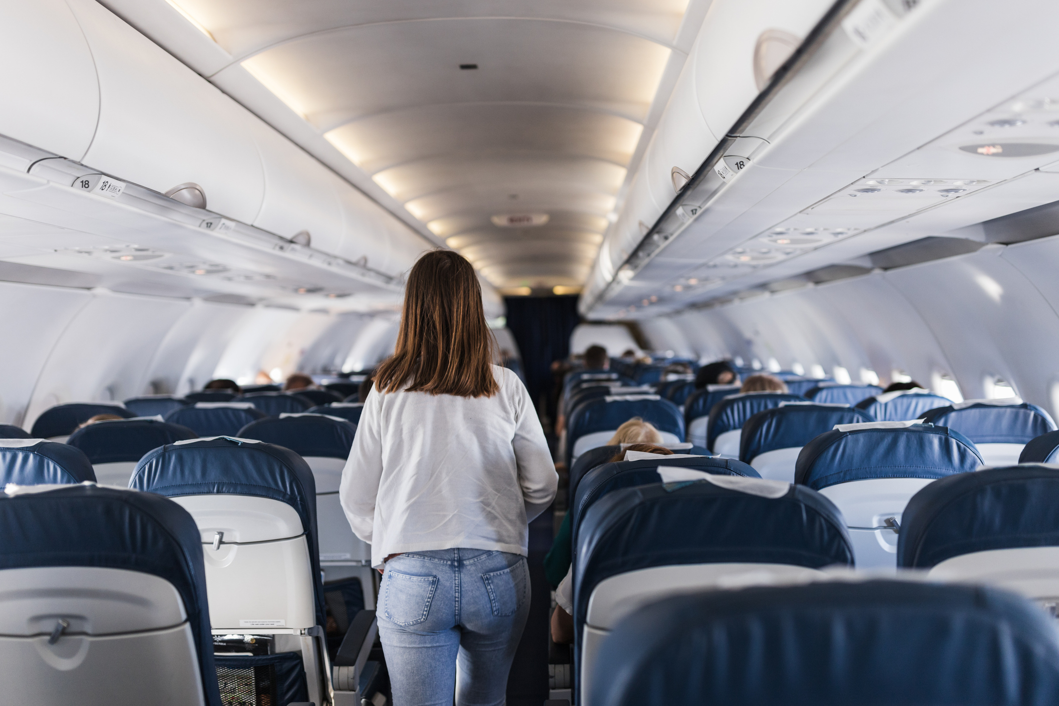 A woman in a white sweater and jeans walks down the aisle of an airplane cabin with several passengers seated