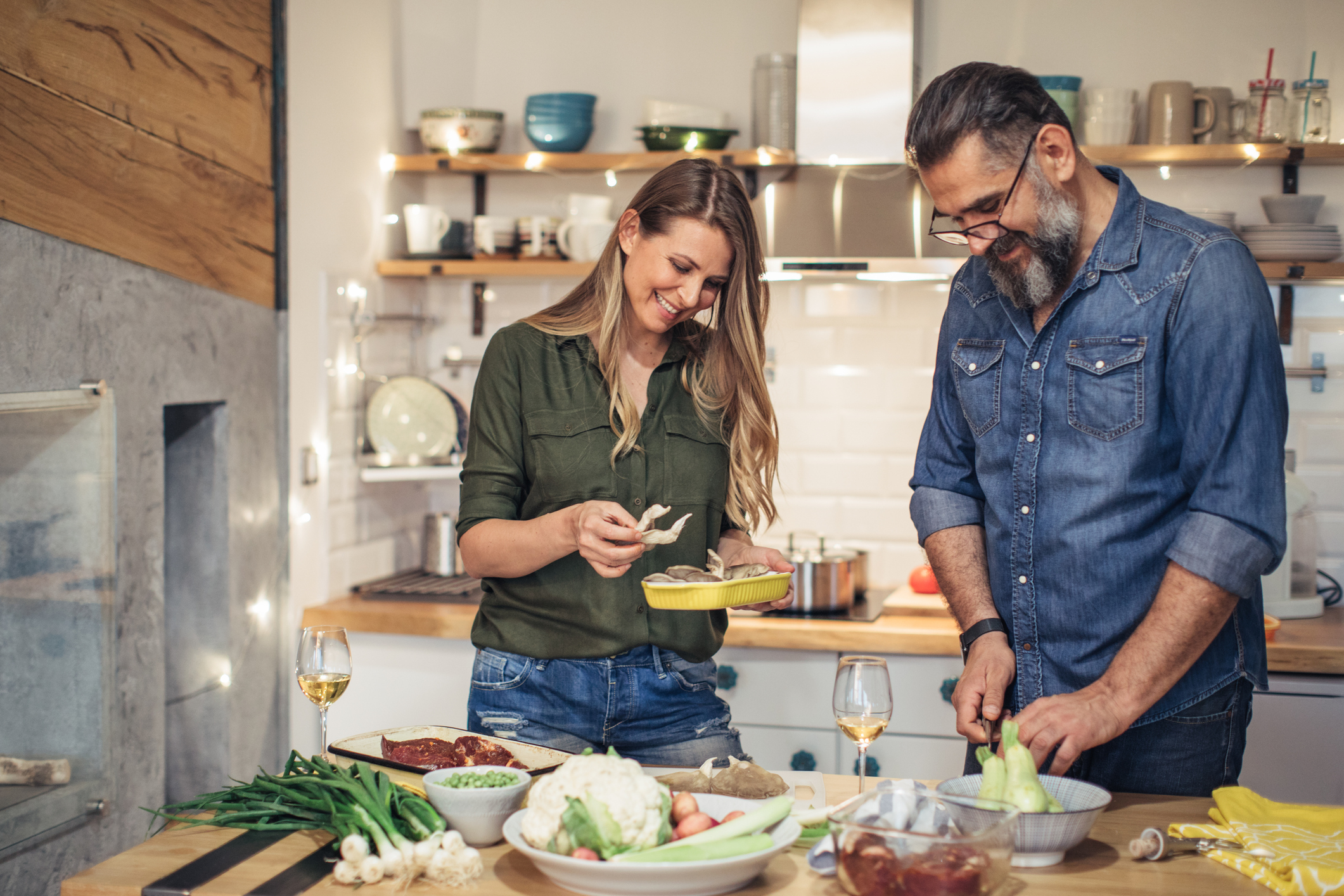 A couple, a woman and a bearded man, are happily preparing a meal together in a modern kitchen with various vegetables and cooking ingredients on the counter