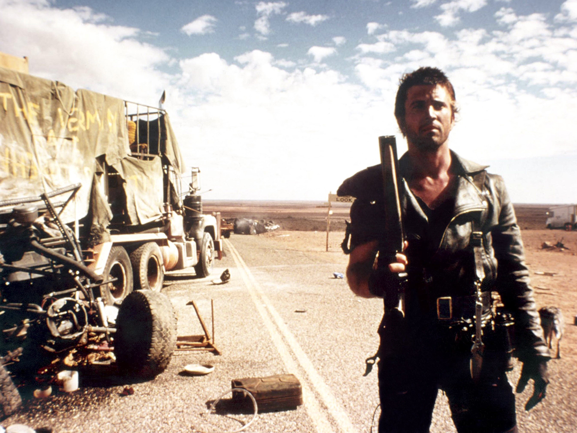 Mel Gibson como Mad Max, sostiene un arma en una carretera desierta. Detrás, un camión dañado y escombros. El paisaje es árido
