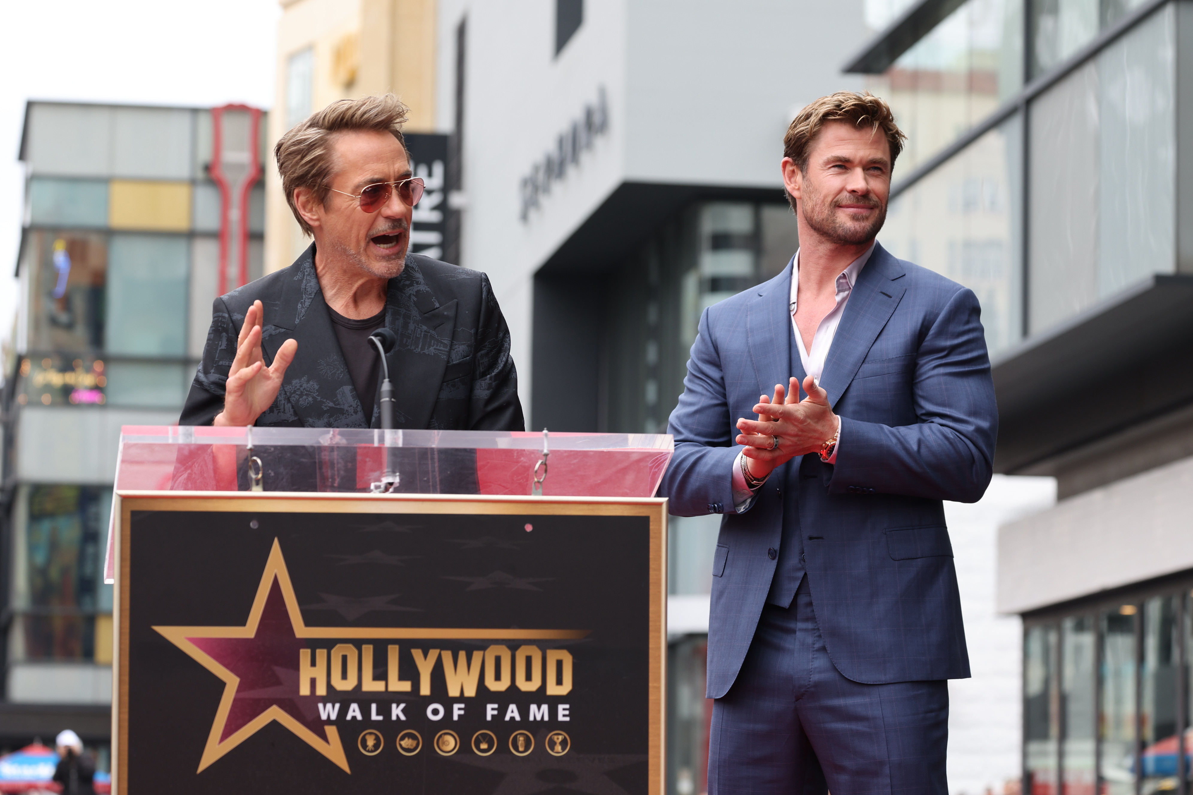 Robert Downey Jr., in a dark suit and sunglasses, speaks at a Hollywood Walk of Fame event while Chris Hemsworth, in a blue suit, stands beside him clapping